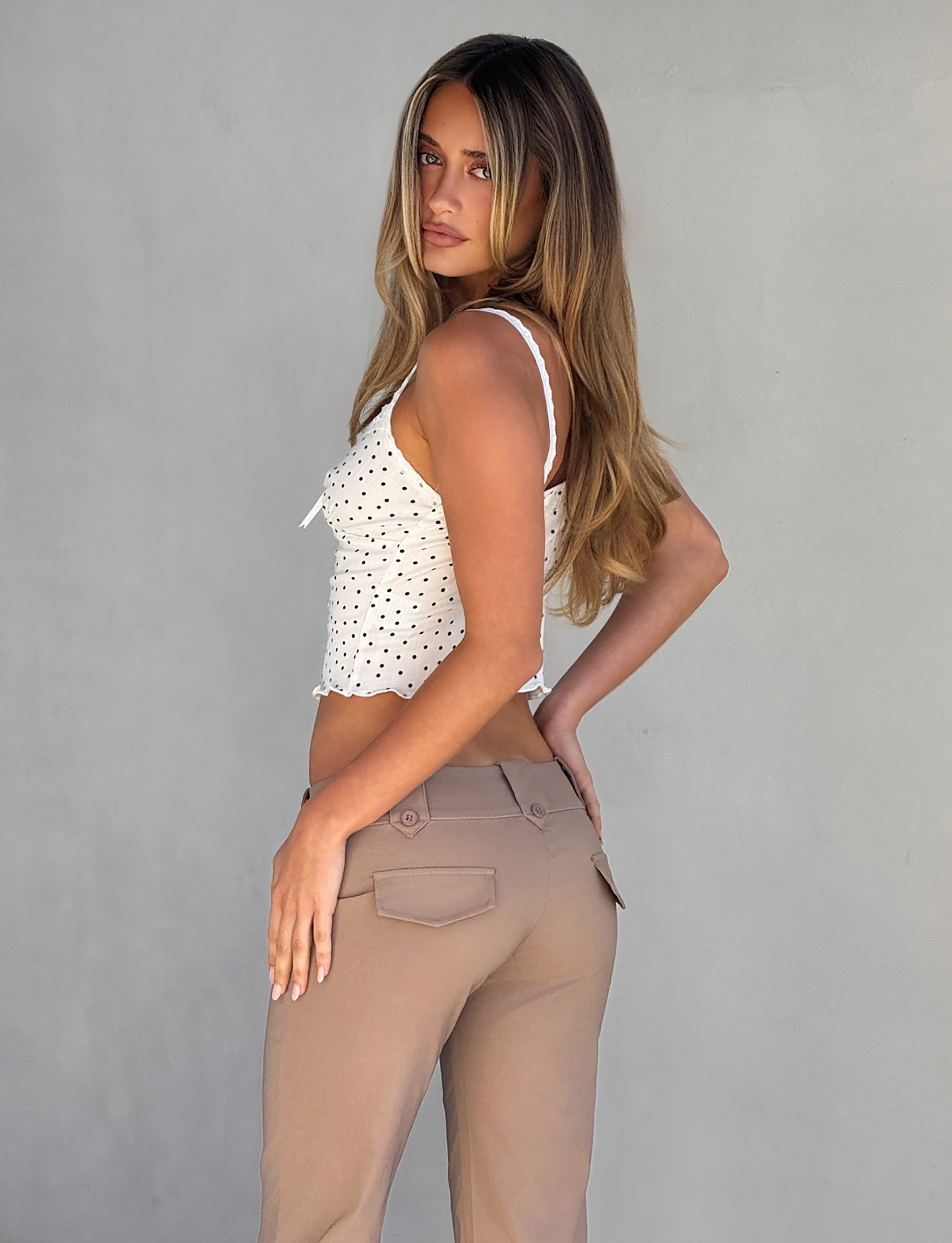 A woman with long, light brown hair stands sideways against a plain gray background, looking over her shoulder at the camera in the BAILEE CAMI TOP - WHITE POLKA by TOP ONE LADYS FASHION PTY LTD, paired with high-waisted beige pants.