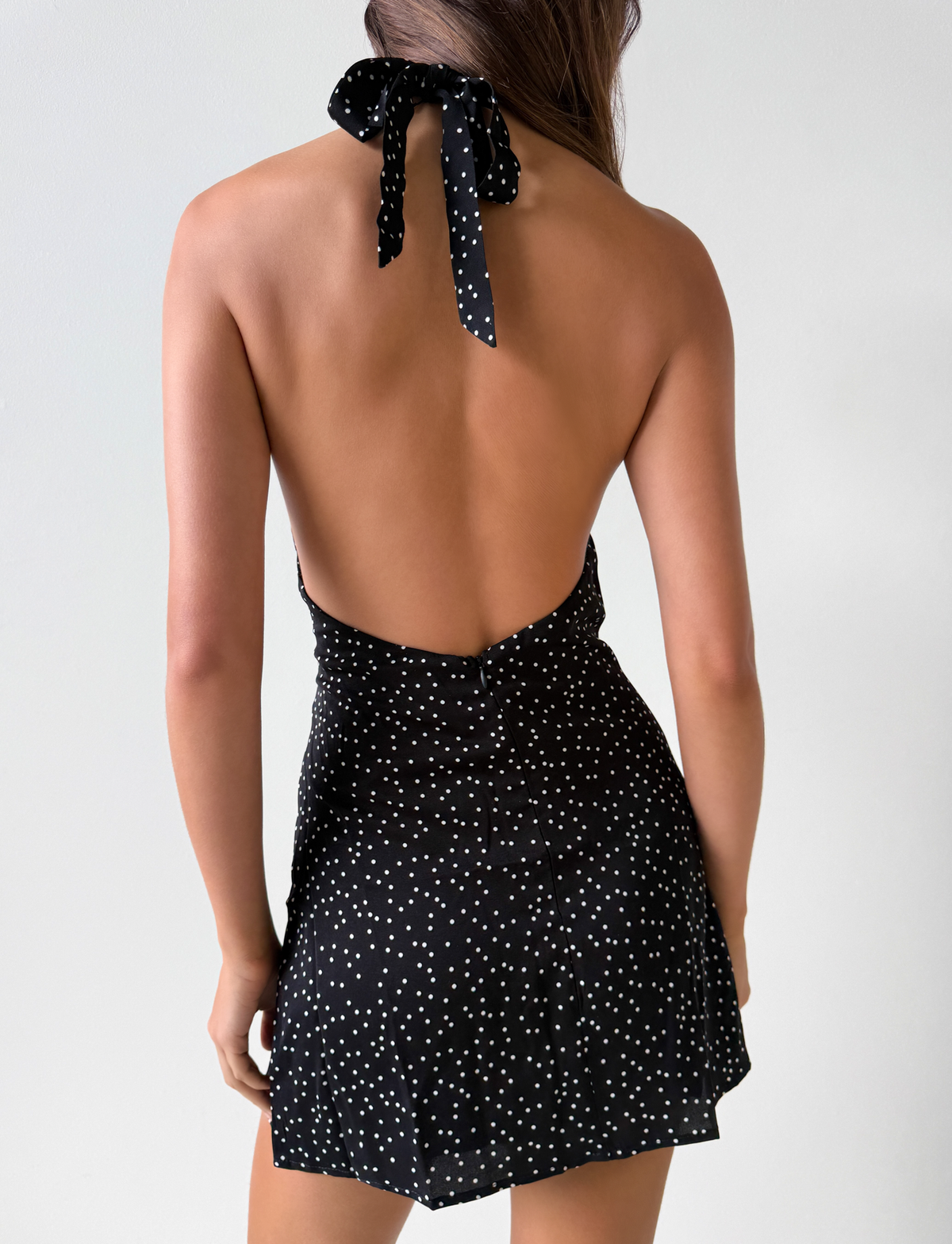 A woman faces away in the DYLAN HALTER MINI DRESS - BLACK POLKA by TOP ONE LADYS FASHION PTY LTD, featuring a black halter mini design with white polka dots and open back, set against a plain white background. Ideal for wholesale purchases.