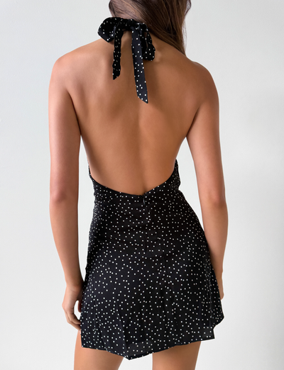 A woman faces away in the DYLAN HALTER MINI DRESS - BLACK POLKA by TOP ONE LADYS FASHION PTY LTD, featuring a black halter mini design with white polka dots and open back, set against a plain white background. Ideal for wholesale purchases.