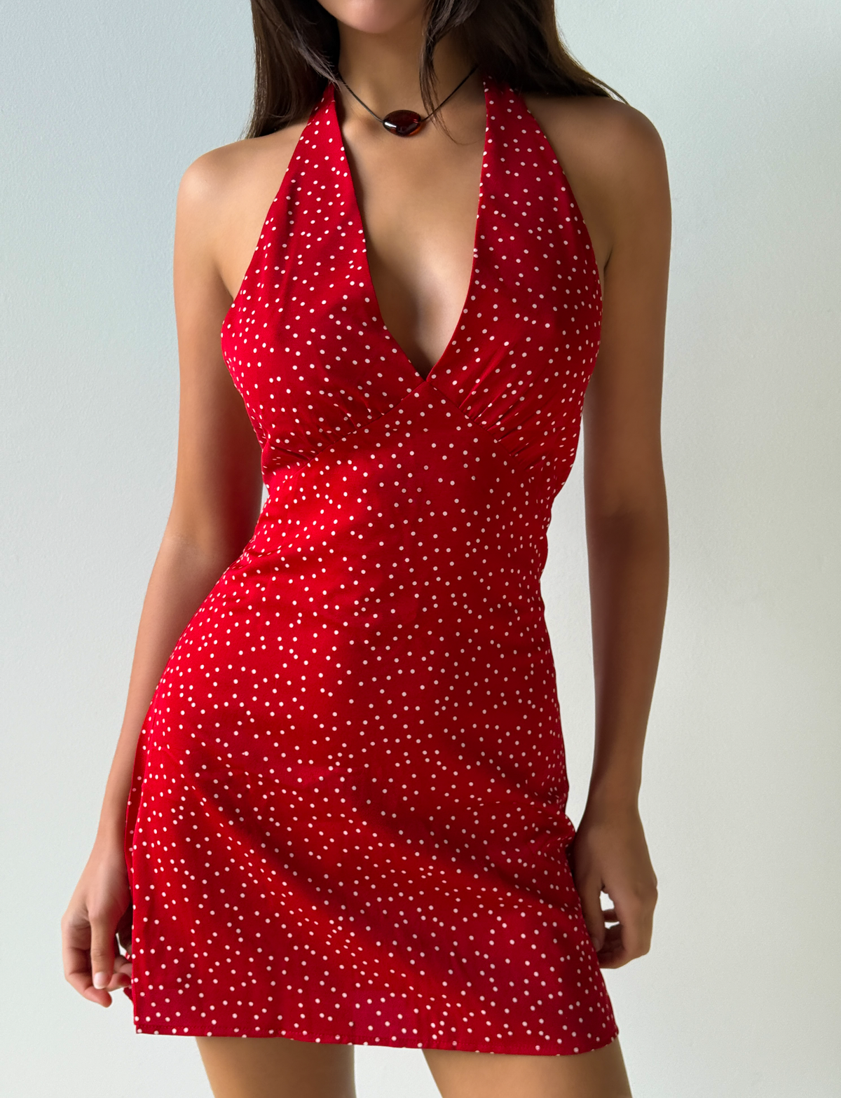 A woman stands against a plain light-colored background wearing the Tiger Mist CL - DYLAN HALTER MINI DRESS, a sleeveless red halter-neck mini dress with white polka dots.