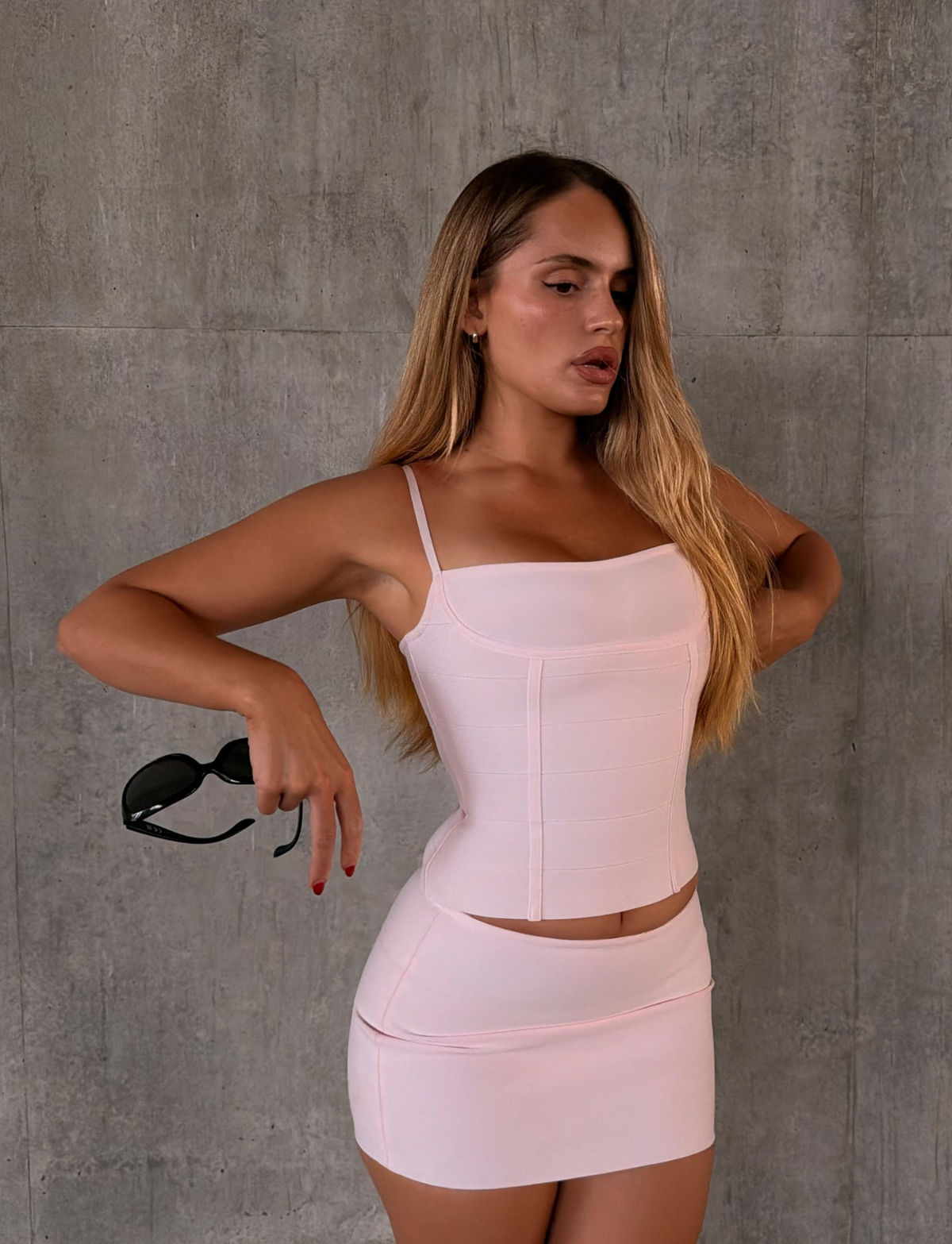 A woman with long hair poses against a concrete wall, wearing a light pink fitted top and the TAHNIA BANDAGE MINI SKIRT in Ballet Pink from TIGERMIST, which accentuates her silhouette. She holds black sunglasses in one hand and looks to the side with a neutral expression.
