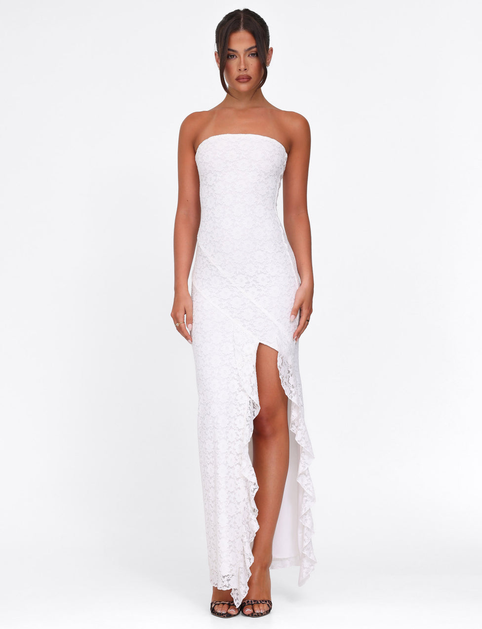 A woman, with tied-back dark hair, stands against a plain white background wearing the TIGERMIST THEO LACE MAXI DRESS - WHITE, a strapless bodycon featuring ruffle details and a high slit, complemented by black open-toed heels.