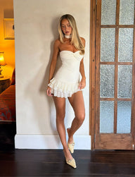 A woman with long blonde hair wears the LYRA MINI DRESS in cream by TIGERMIST, featuring a corset bodice and lace details, paired with cream heels as she stands indoors by a frosted glass door on a dark wood floor, illuminated by soft lighting from a nearby lamp.