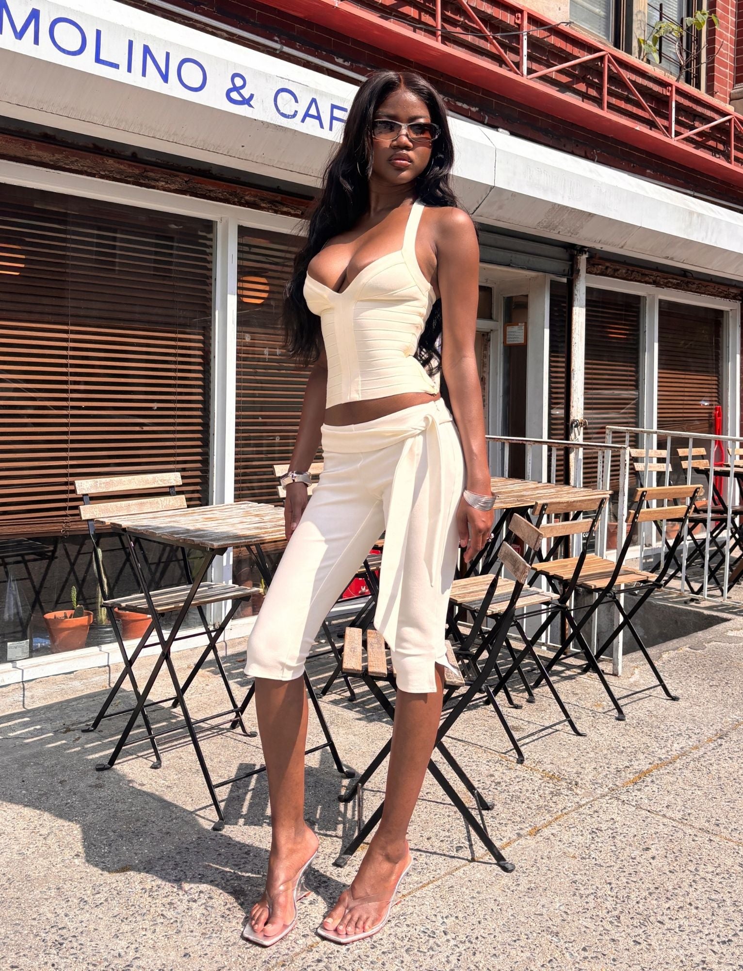 A woman stands confidently on a sunny sidewalk outside a cafe with wooden chairs and tables, dressed in a stylish cream-colored ensemble featuring the TIGERMIST PILAR CAPRI - WHITE pants, which showcase a slim leg and removable waist tie.