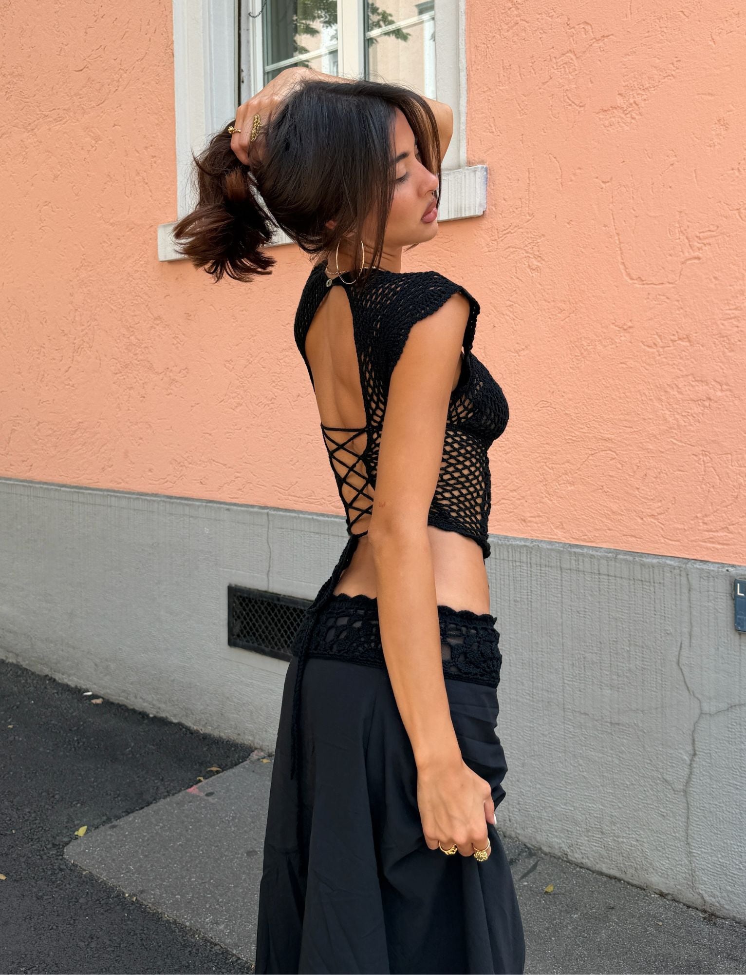 A woman stands outside against a peach wall, wearing the TIGERMIST SASHA CROCHET TOP in black—a floral crochet design featuring an open lace-up back—paired with a matching black skirt. Channeling effortless resort style, she poses with one hand in her hair and glances over her shoulder.