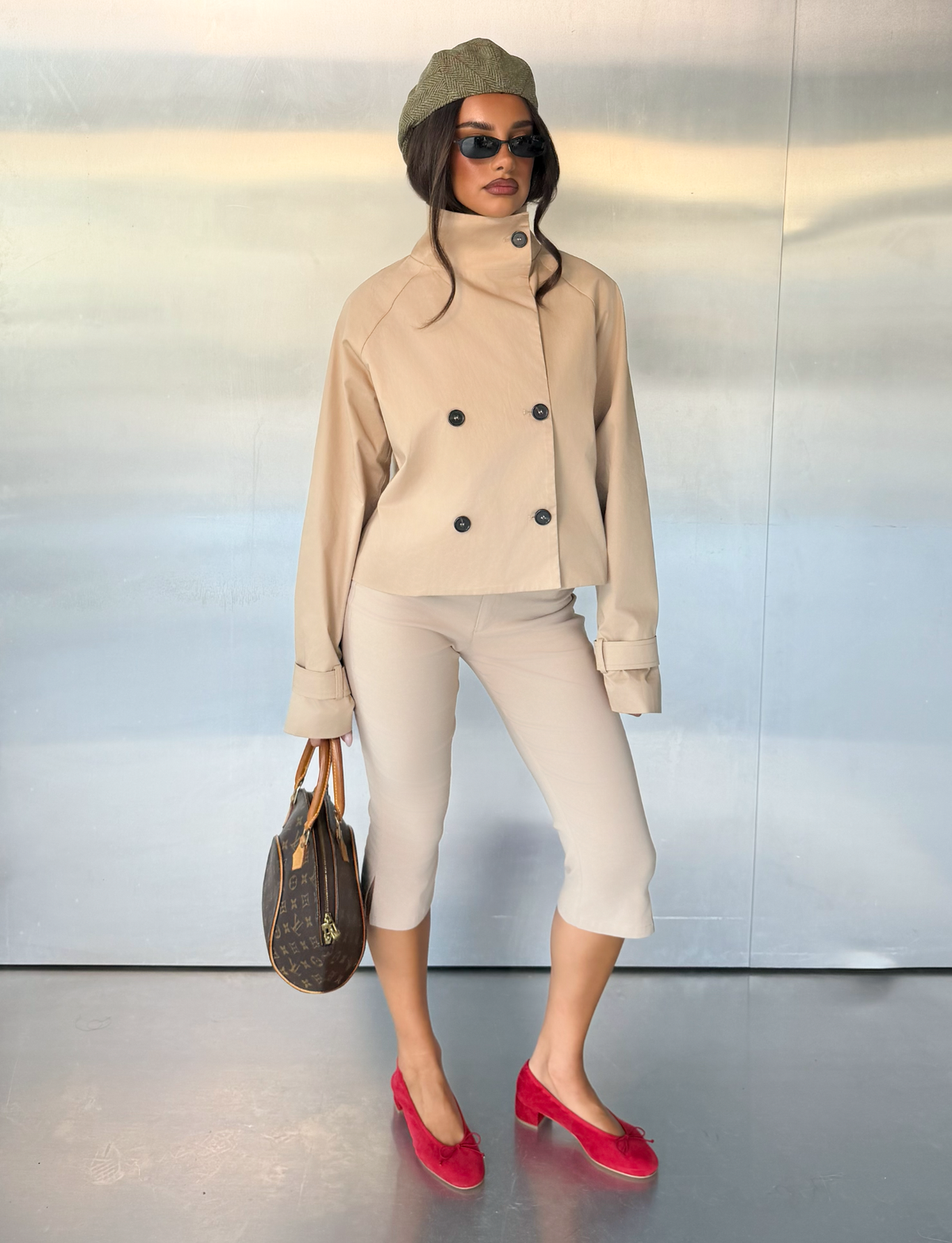 A woman stands against a metallic wall holding a brown handbag, wearing the TIGERMIST HAYLEY CROPPED TRENCH JACKET in khaki, capri pants, red shoes, sunglasses, and a green beret.