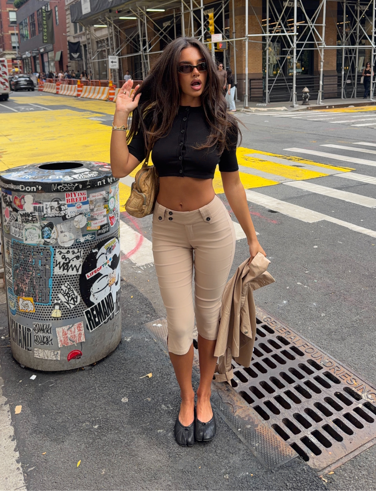 A woman with long hair and sunglasses stands on a city street, wearing a black crop top, black shoes, and the TIGERMIST ARIA CAPRI - KHAKI tailored pants. She holds a tan jacket and poses near a decorated trash can by a construction site.