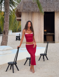 A woman barefoot on sand poses near a thatched hut, palm trees, and chairs, wearing the TIGERMIST CALINE MIDI SKIRT in red and a sheer mesh crop top.