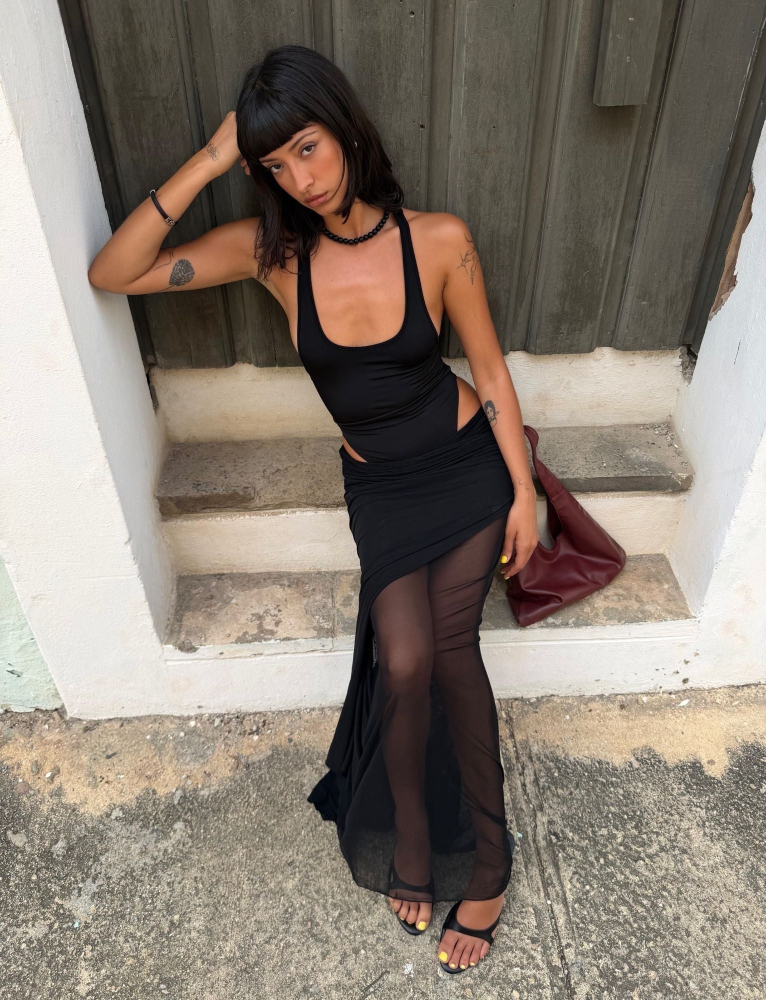 A woman with straight dark hair sits on concrete steps, wearing the TIGERMIST LILIANA-SUMMER MAXI DRESS in black with a sheer mesh skirt, paired with black heels and holding a dark red handbag as she gazes thoughtfully to the side.