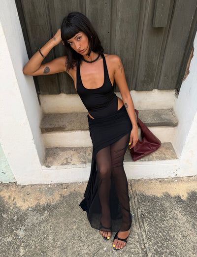 A woman with straight dark hair sits on concrete steps, wearing the TIGERMIST LILIANA-SUMMER MAXI DRESS in black with a sheer mesh skirt, paired with black heels and holding a dark red handbag as she gazes thoughtfully to the side.