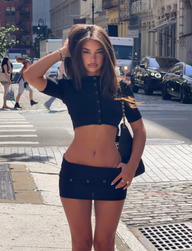 A woman in a black crop top and skirt stands on a city street, posing with one hand in her hair. She layers her look with the TIGERMIST ELSIE CARDIGAN in black—a super soft knit cropped cardigan featuring faux shell buttons. Cars and pedestrians are visible in the background on a sunny day.