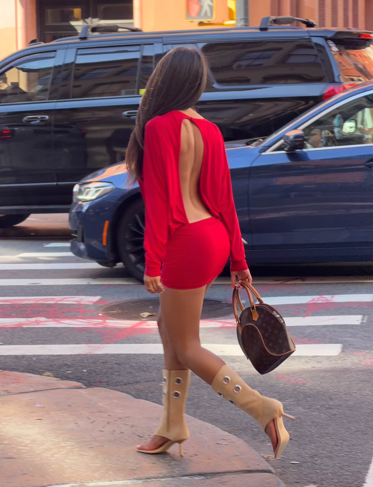 A woman wearing the TIGERMIST BIANCA MINI DRESS in red, styled with tan knee-high boots and carrying a brown designer handbag, walks across a city street with cars and buildings in the background.