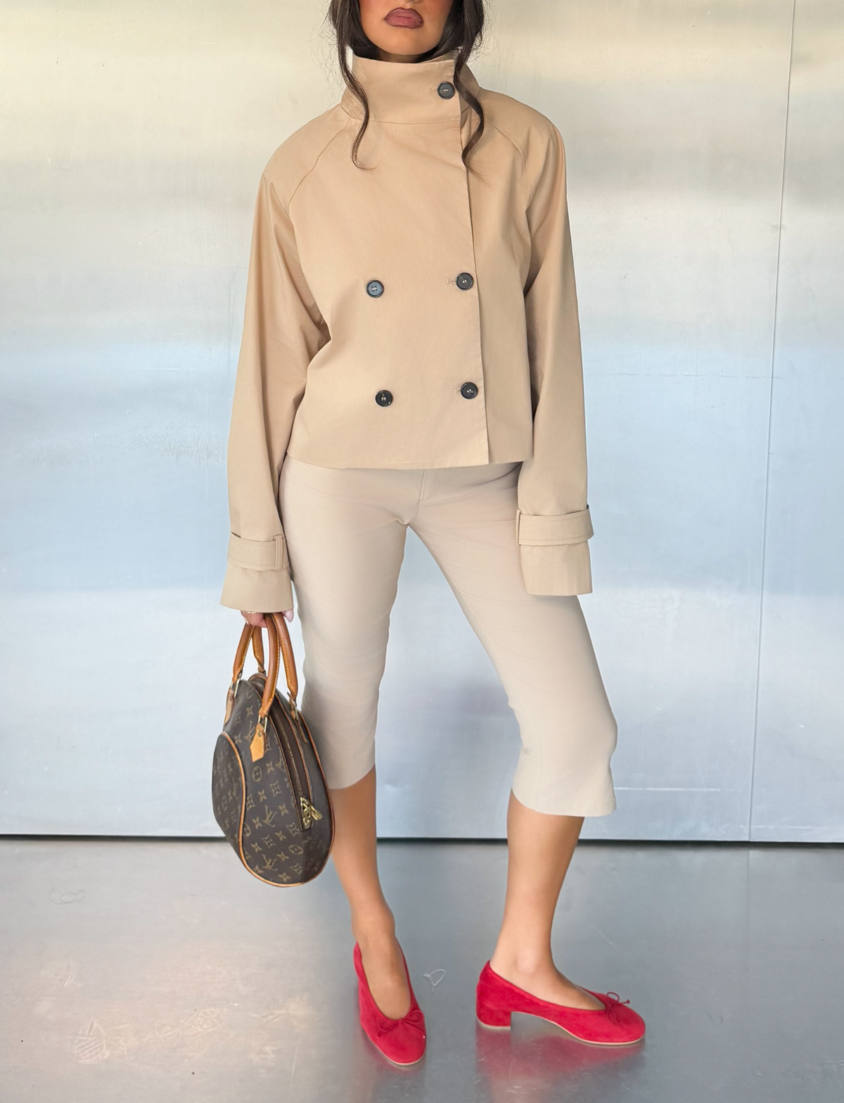 A woman stands in a relaxed pose against a plain, metallic background, wearing the TIGERMIST HAYLEY CROPPED TRENCH JACKET in khaki paired with beige cropped pants and red flats. She holds a designer handbag, with her hair partially visible.
