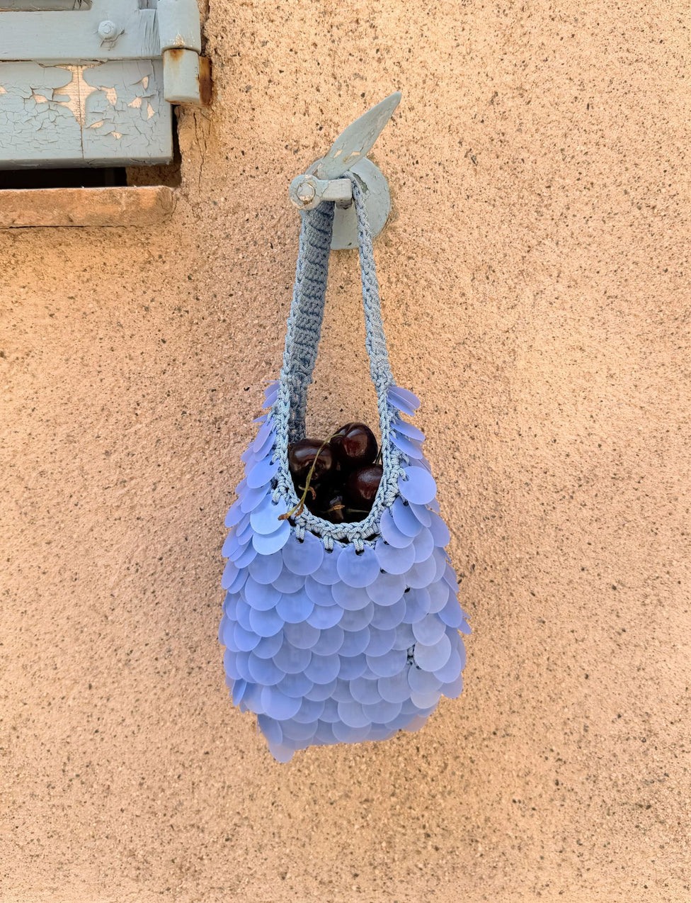 The CLEO BAG in Bright Blue by TIGERMIST, featuring scale-like crochet decorations, hangs on a beige wall and holds several dark cherries. This eye-catching piece, reminiscent of a sequin crochet bag, is set below a weathered blue wooden shutter in the upper left corner.
