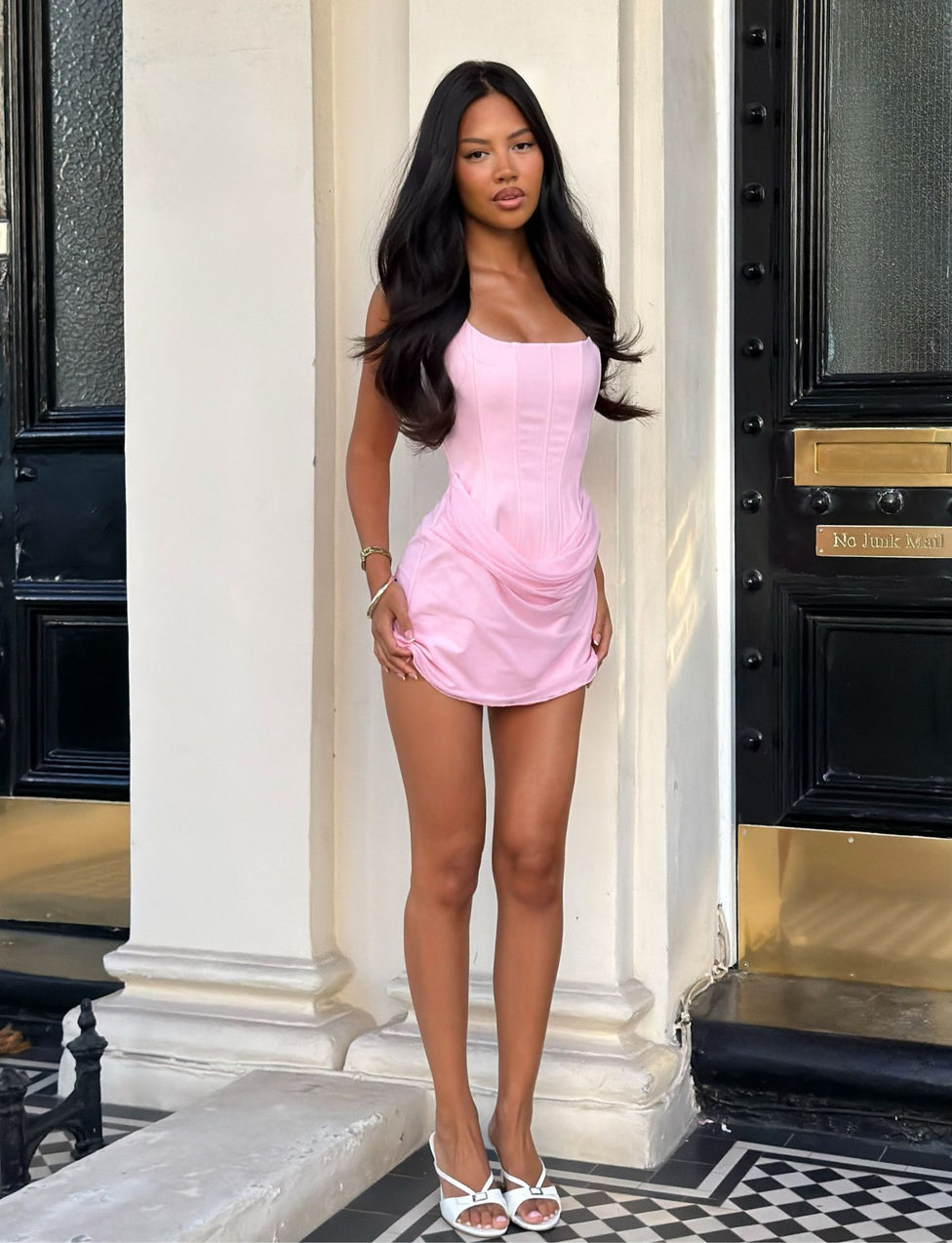 A woman with long dark hair stands on a tiled entryway in front of a building with black doors and gold accents, wearing the TIGERMIST LYZA MINI DRESS in baby pink and white sandals.
