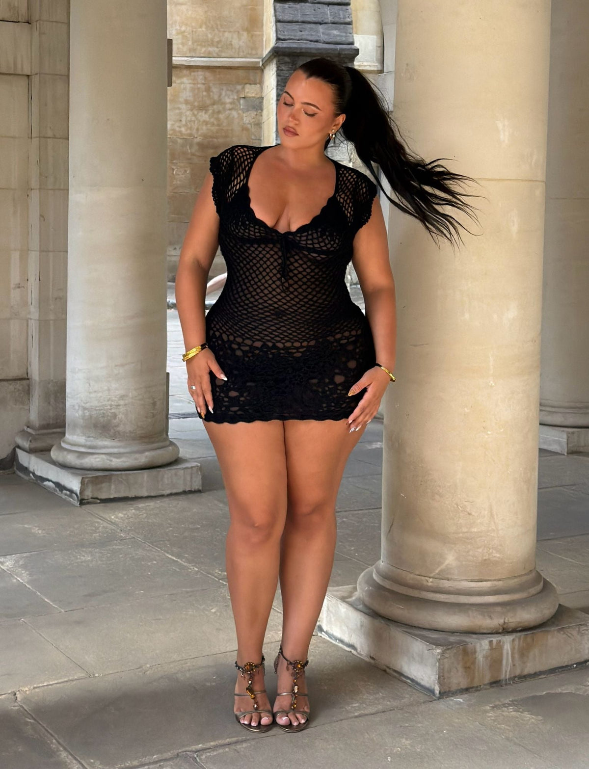 A woman wearing the TIGERMIST SASHA CROCHET MINI DRESS in black and sandals stands between stone columns, gazing down with one hand resting on her thigh. Her long hair is pulled back, flowing in the wind. The backdrop showcases tall pillars and stone walls.