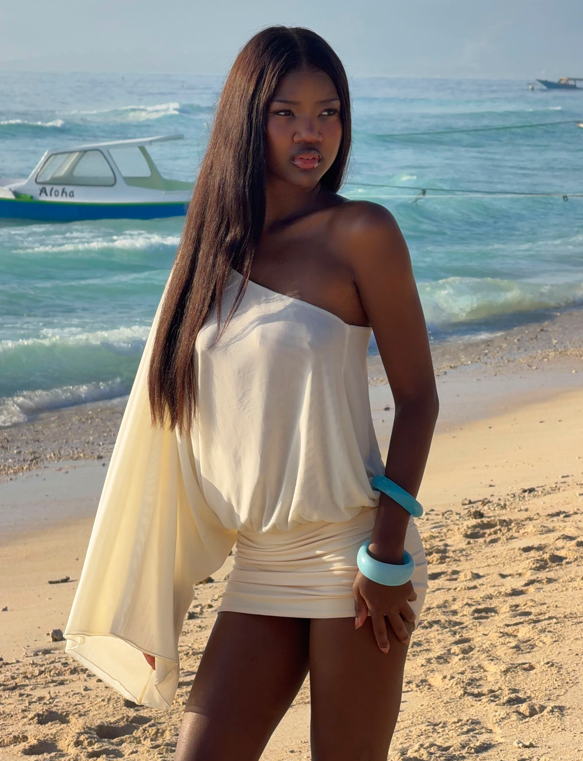 A woman in a white, one-shoulder dress stands on a sandy beach, wearing the TIGERMIST MIKAH BANGLE SET - AQUA as the ocean and a boat create a serene background.