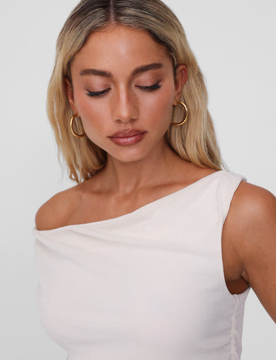 A long-haired blonde woman wears TIGERMIST's PIPER EARRING in gold and an asymmetrical one-shoulder white top. She gazes down neutrally against a plain gray background.