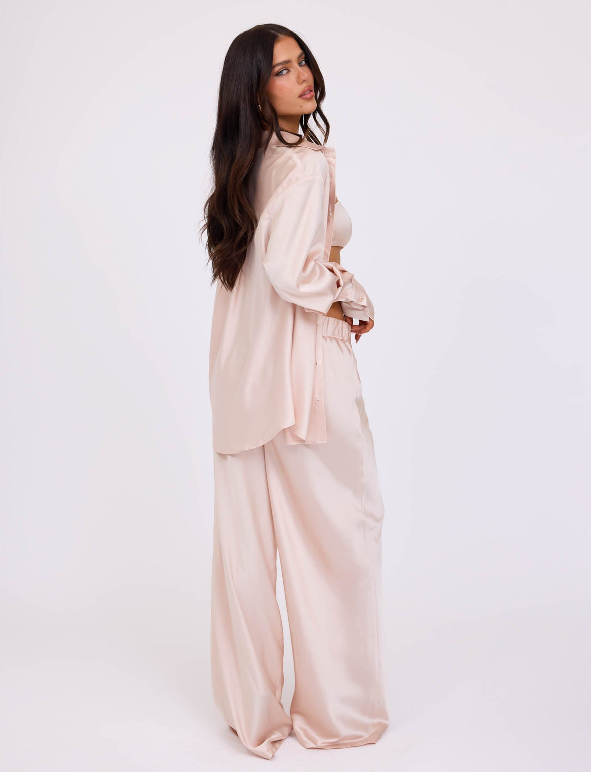 A woman with long dark hair stands in profile, dressed in a light pink, loose-fitting satin outfit from TIGERMIST. She wears the ALINA SHIRT - WHITE : IVORY, a long-sleeved button-up shirt over matching wide-leg pants. The plain white background emphasizes her stylish and relaxed ensemble.