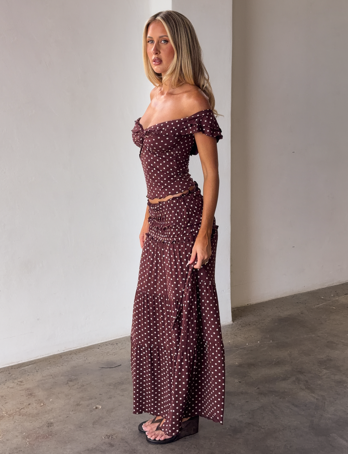 A woman with long blonde hair stands indoors against a white wall, wearing the TIGERMIST DENALI MAXI SKIRT - BROWN DOT, an off-the-shoulder top, and black platform sandals, looking slightly to her left.