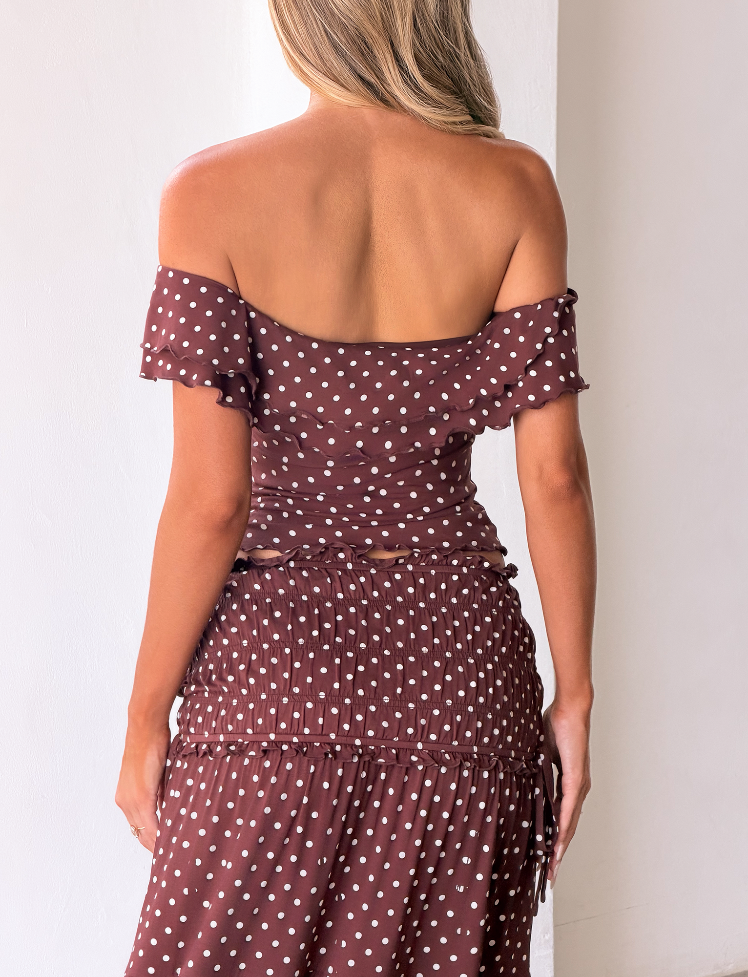 A woman with long blonde hair faces away, wearing the TIGERMIST DENALI TOP in brown dot—a polka dot, off-the-shoulder top with ruffled sleeves—contrasting against a plain light background.