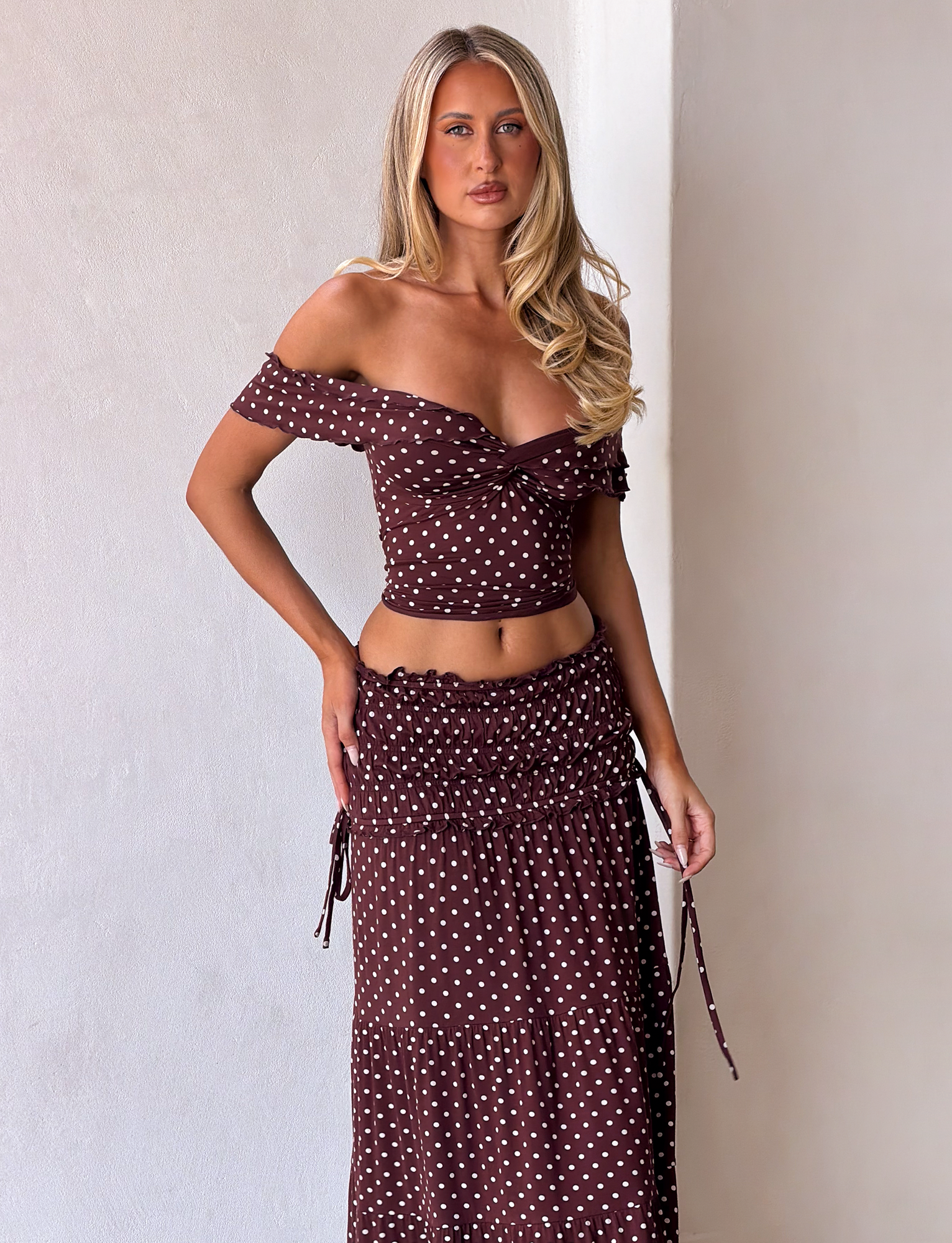 A woman with long blonde hair poses against a light wall, wearing the TIGERMIST DENALI MAXI SKIRT - BROWN DOT and a brown off-shoulder crop top, with one hand on her hip and a relaxed expression.