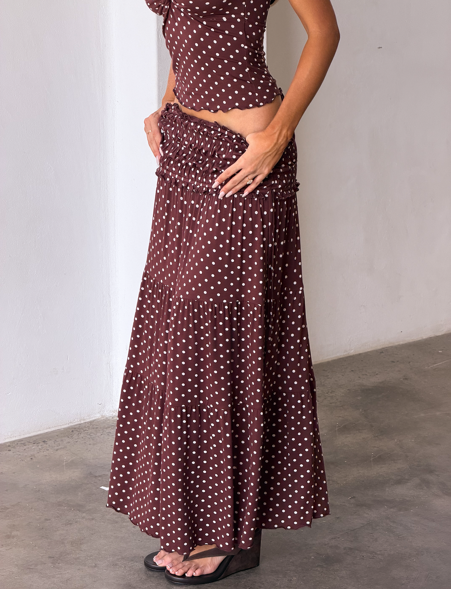 A woman in the TIGERMIST DENALI MAXI SKIRT - BROWN DOT and matching top stands against a white wall, hands on hips and wearing black platform sandals with white polka dots.