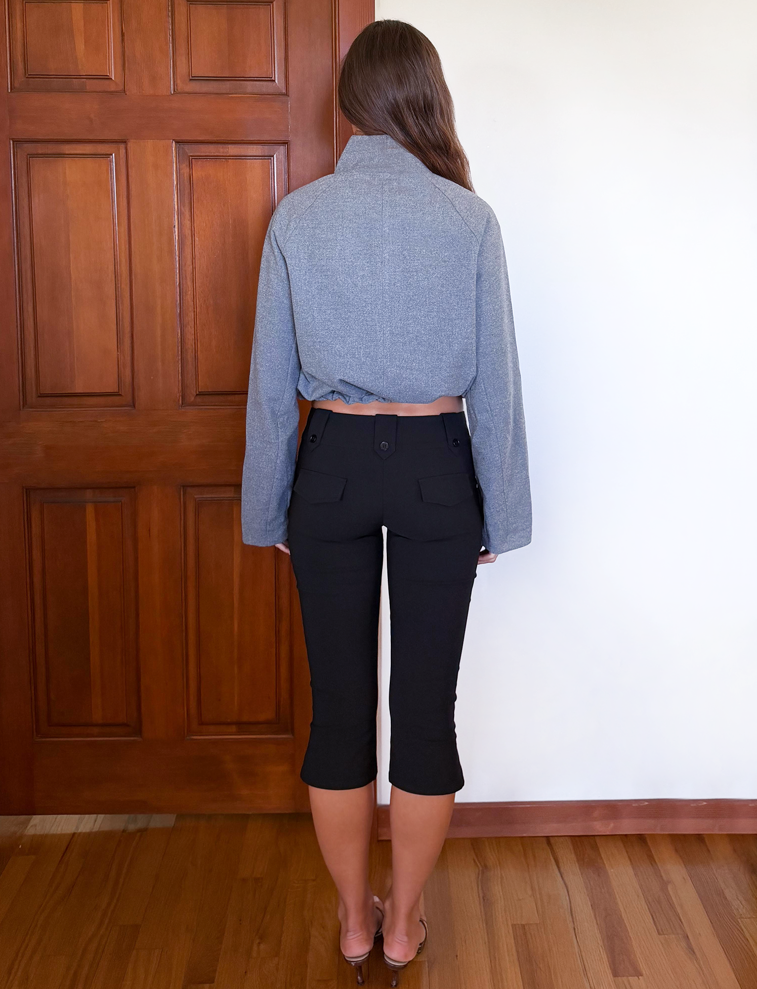A person with long brown hair stands indoors, facing away, wearing the TIGERMIST LENNEX JACKET in heather charcoal and black cropped pants on a wooden floor near a wooden door and white wall.