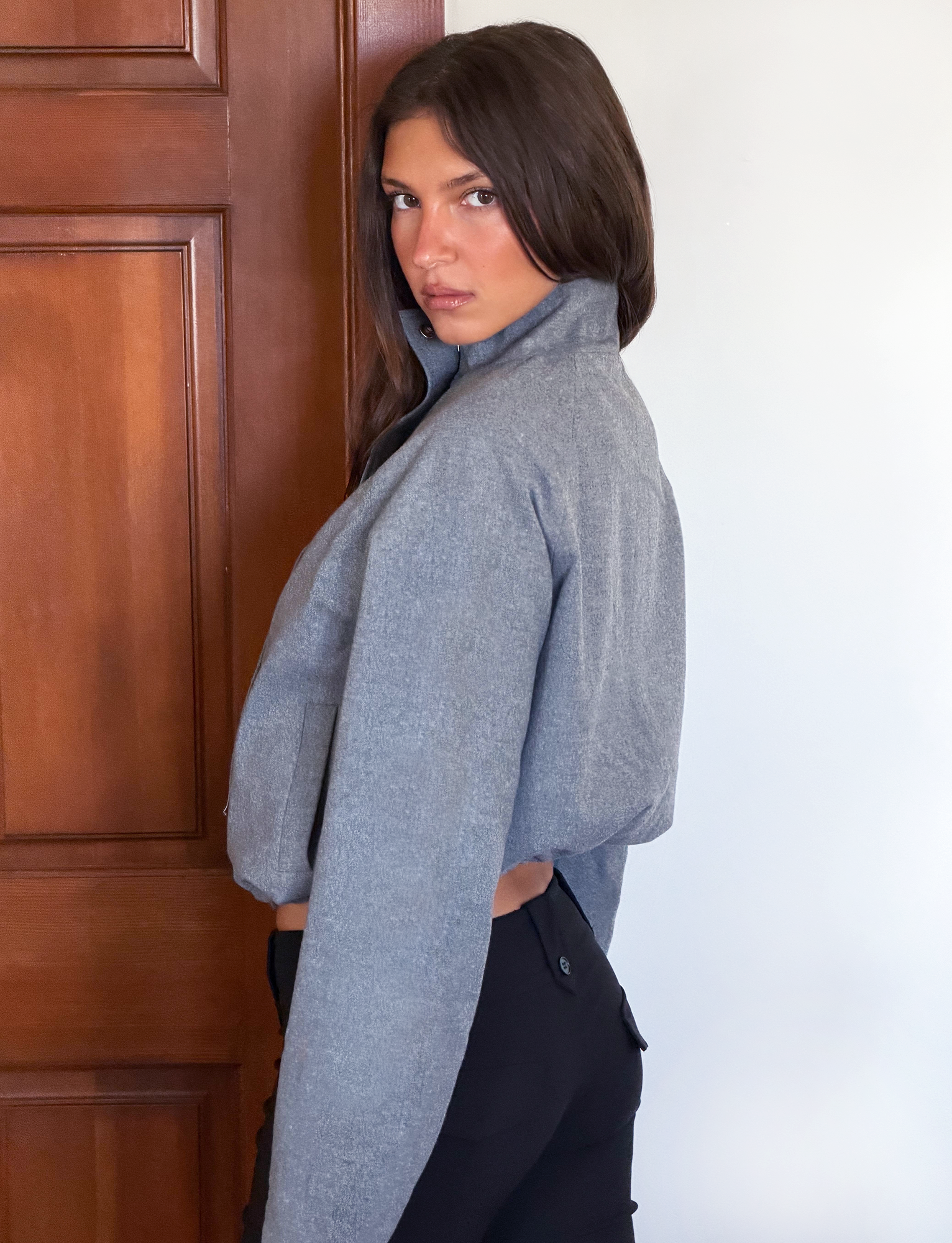 A woman with long brown hair stands sideways, looking over her shoulder at the camera. She wears the TIGERMIST LENNEX JACKET in Heather Charcoal and black pants, with a wooden door and white wall behind her.