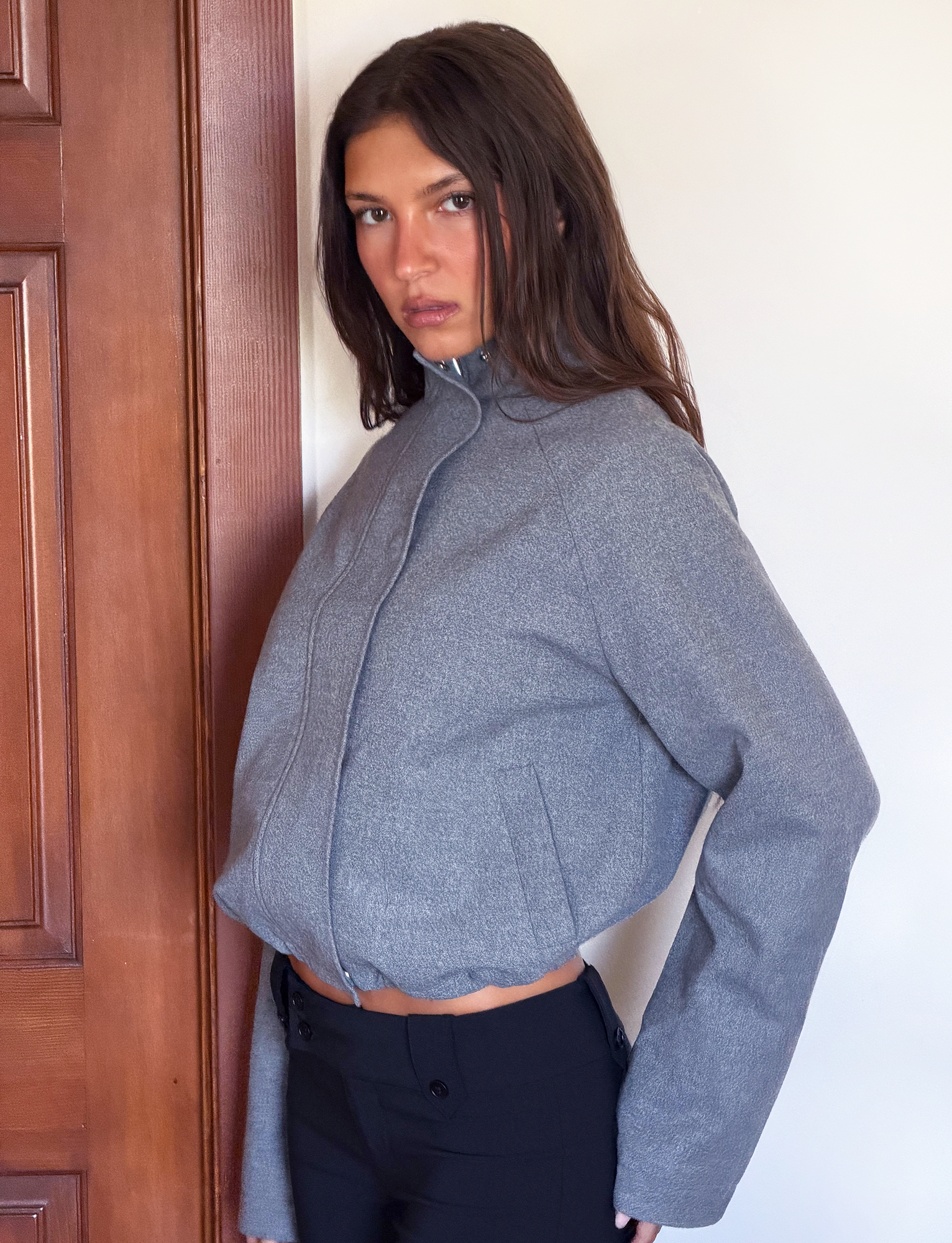 A person with long brown hair wears the TIGERMIST LENNEX JACKET in Heather Charcoal, styled oversized with a bungee drawstring hem and black pants, standing indoors against a white wall and wooden door, gazing seriously at the camera.