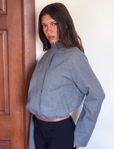 A person with long brown hair wears the TIGERMIST LENNEX JACKET in Heather Charcoal, styled oversized with a bungee drawstring hem and black pants, standing indoors against a white wall and wooden door, gazing seriously at the camera.