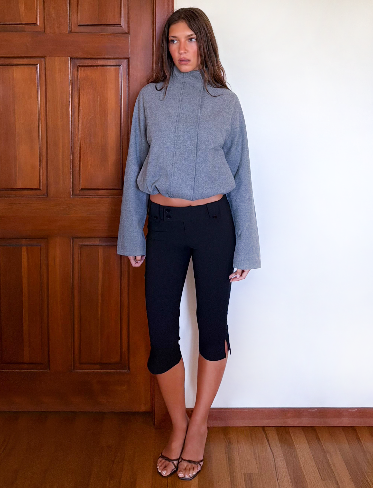 A woman with long brown hair and a neutral expression stands indoors against a white wall and wooden door, wearing the TIGERMIST LENNEX JACKET in heather charcoal, styled with black cropped pants and open-toe heels.