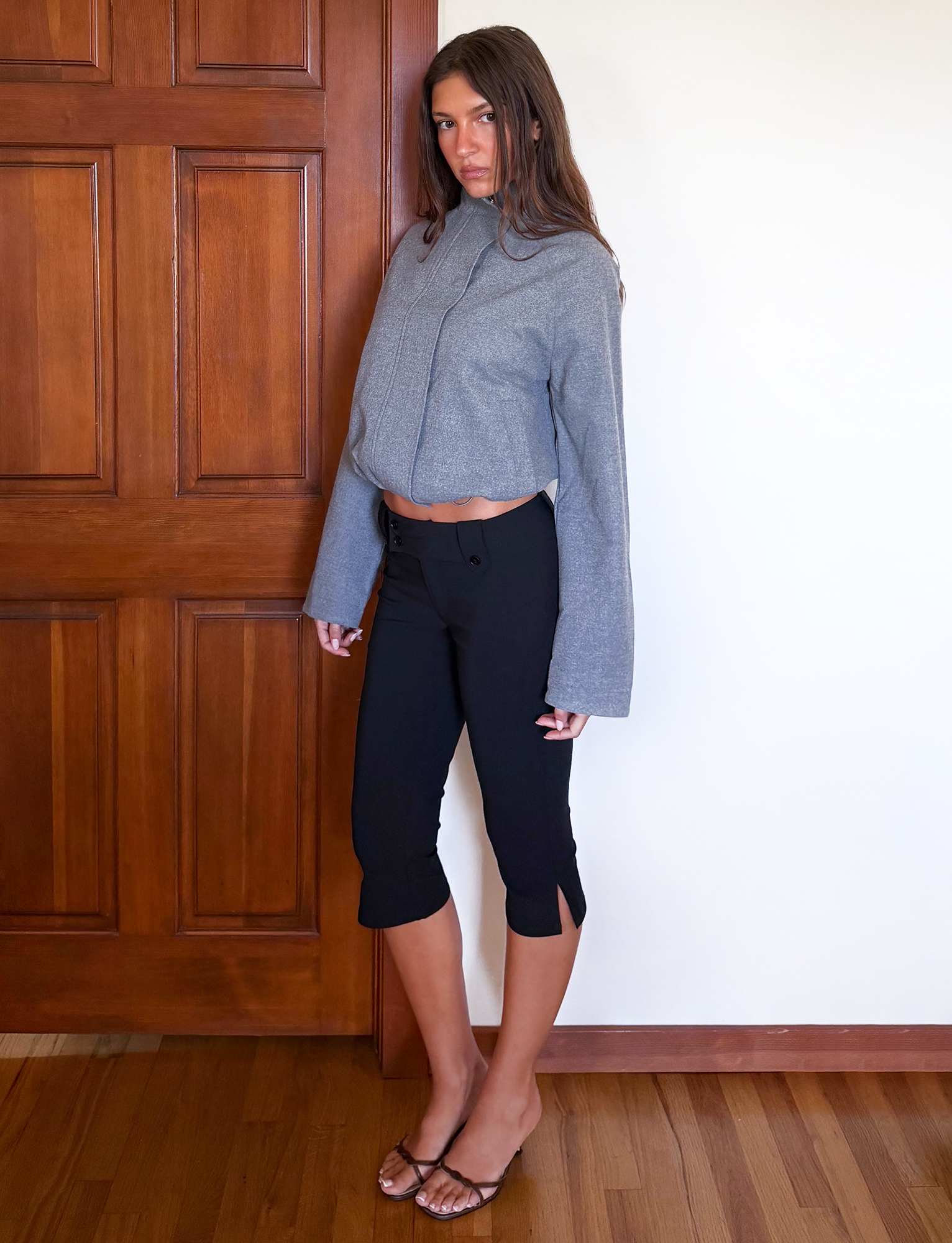A woman stands indoors against a wooden door and white wall, wearing the TIGERMIST LENNEX JACKET in heather charcoal, black cropped pants with side slits, and brown sandals. She looks directly at the camera with a neutral expression.