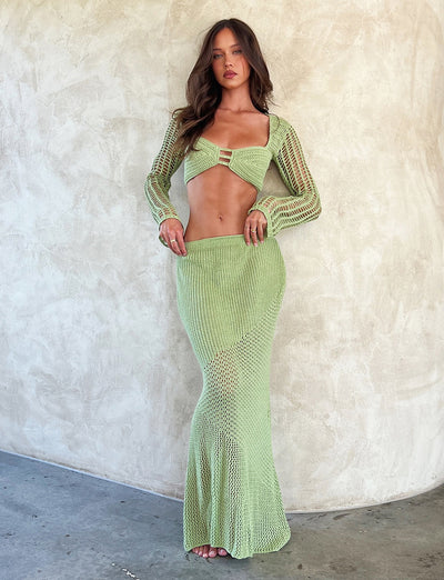 A woman with long brown hair is posing against a plain, textured beige wall. She is wearing the MACAULEY SKIRT - GREEN : PASTEL GREEN from TIGERMIST, featuring a light green knitted two-piece outfit with a cropped long-sleeve top with cut-out details and a matching high-waisted maxi skirt.