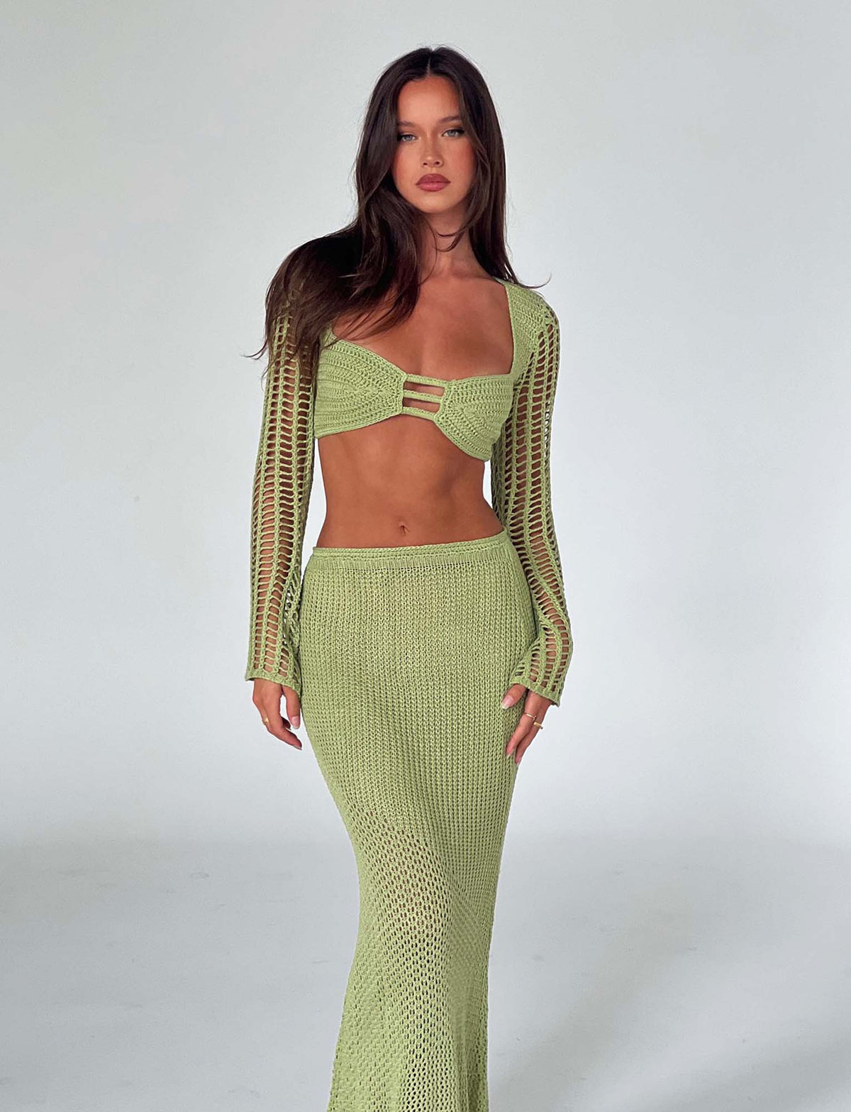A woman with long brown hair stands against a plain white background, wearing a light green MACAULEY TOP - PASTEL GREEN by TIGERMIST. The ensemble consists of a cropped top with long sleeves and a keyhole detail, paired with a matching long, fitted skirt.