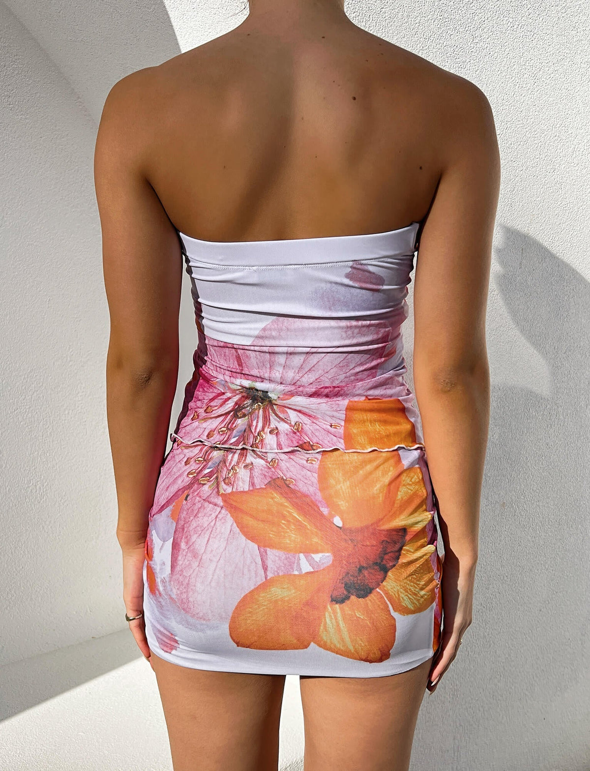 A person is standing with their back to the camera, wearing a strapless white dress decorated with large orange and pink flowers from Tigermist. The background is a plain, light-colored wall.

