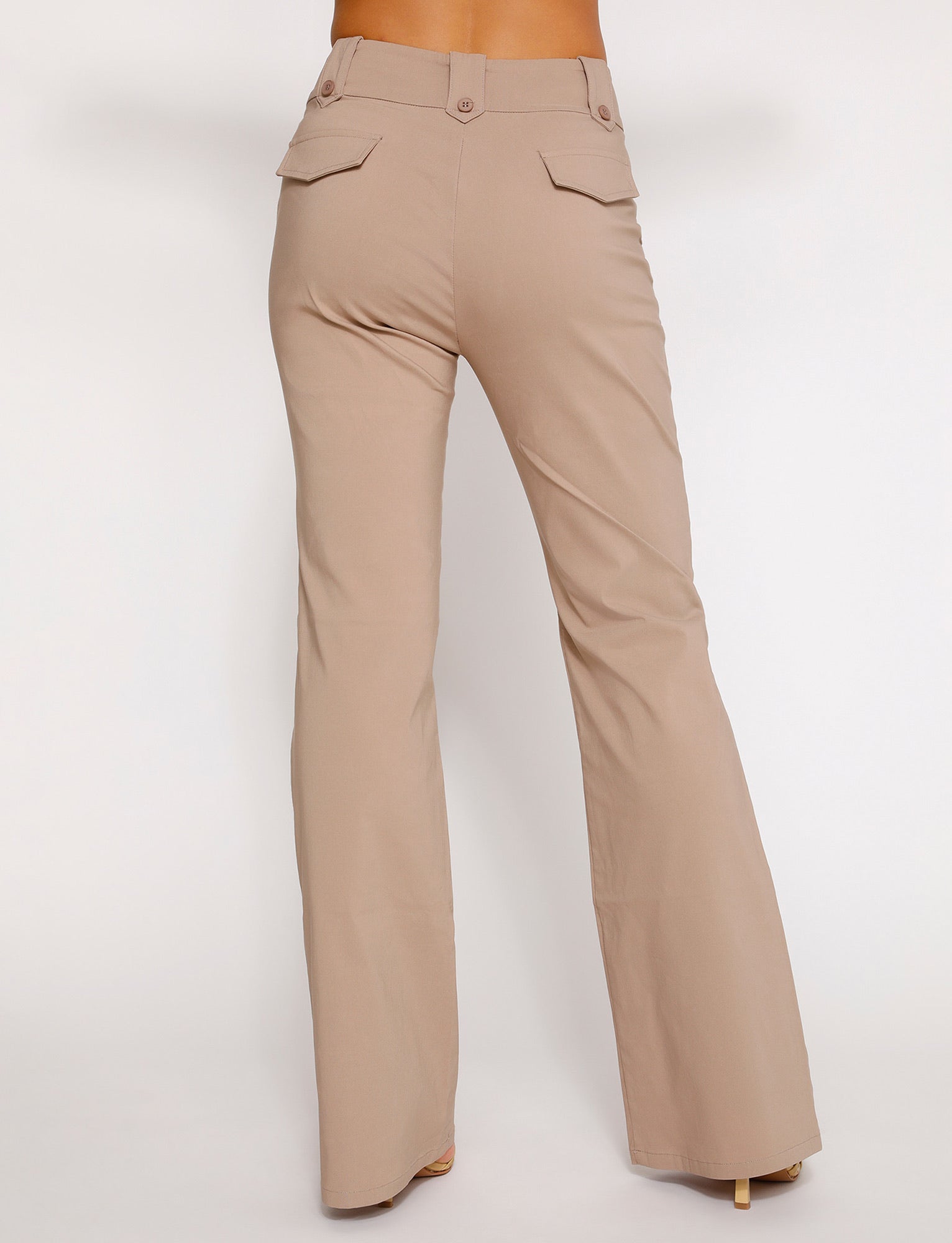 A person wearing the KITTIE HIGH RISE PANT - BEIGE : TAN from TIGERMIST stands with their back facing the camera. The high-waisted, wide-leg pants feature buttoned flap pockets on the back and are made of a smooth fabric. The background is plain white.