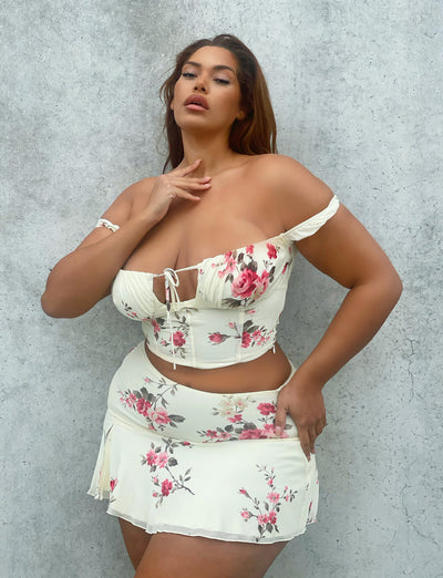 A woman stands confidently in front of a textured gray wall, wearing the TIGERMIST FEYRA TOP in yellow with a macro floral pattern. The top features off-the-shoulder sleeves and a small tie in the front. The matching short skirt is adorned with a similar pink and red floral design. She poses with one hand on her chest.