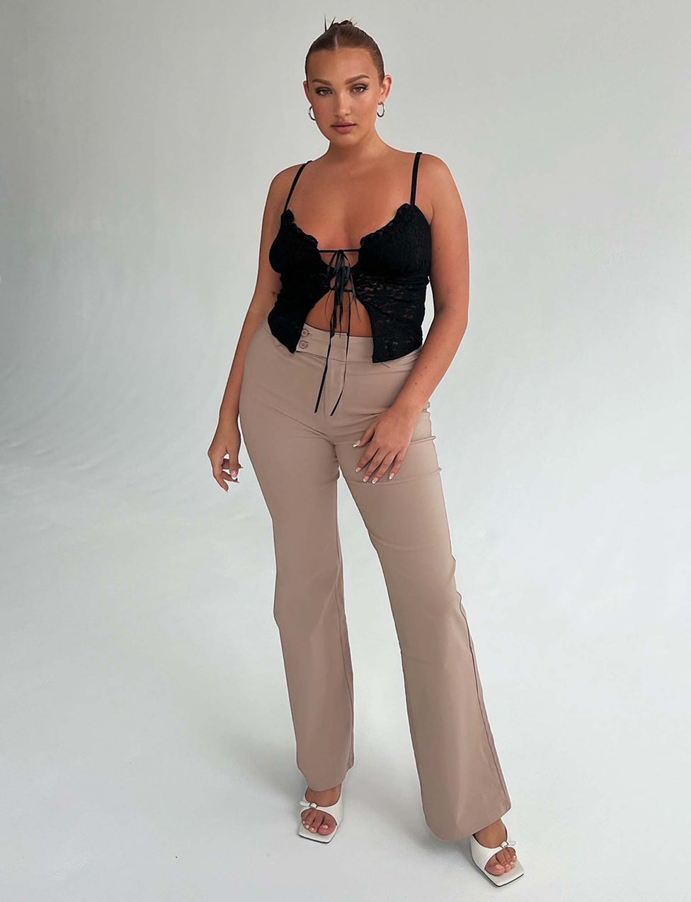 A person stands posed against a white background in an edgy black lace-up top from Tiger Mist, paired with the Kittie High Rise Pant in beige from TIGERMIST. They are also wearing white heeled sandals and large hoop earrings, with their hair styled back while they gaze confidently at the camera.