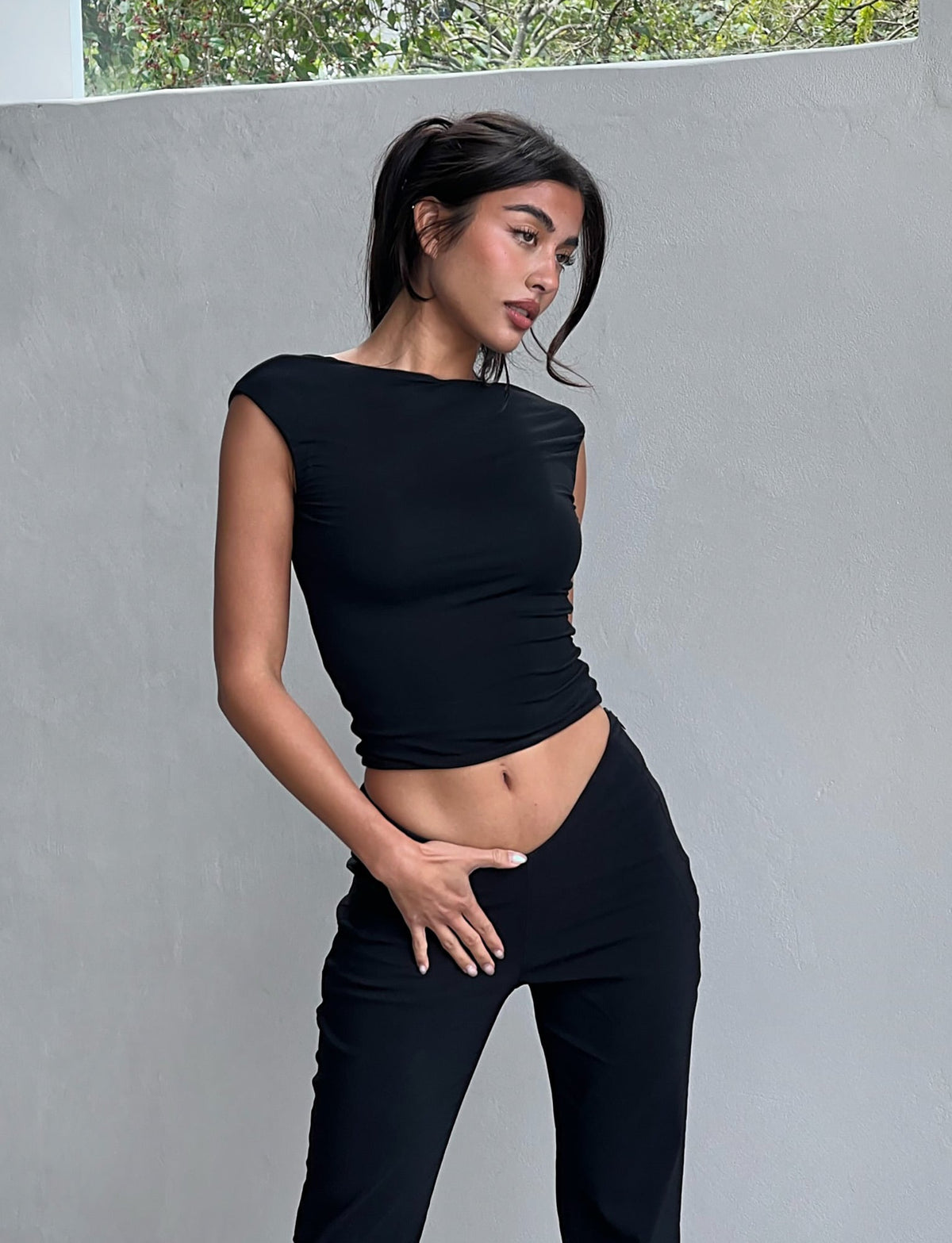 A woman stands against a plain gray wall, gazing to her left. She wears the fitted, sleeveless 'CHRISTY TOP - BLACK' from TIGERMIST with black pants, one hand on her hip and the other resting on her stomach. Her dark hair is pulled back in a casual ponytail, and she appears thoughtful.
