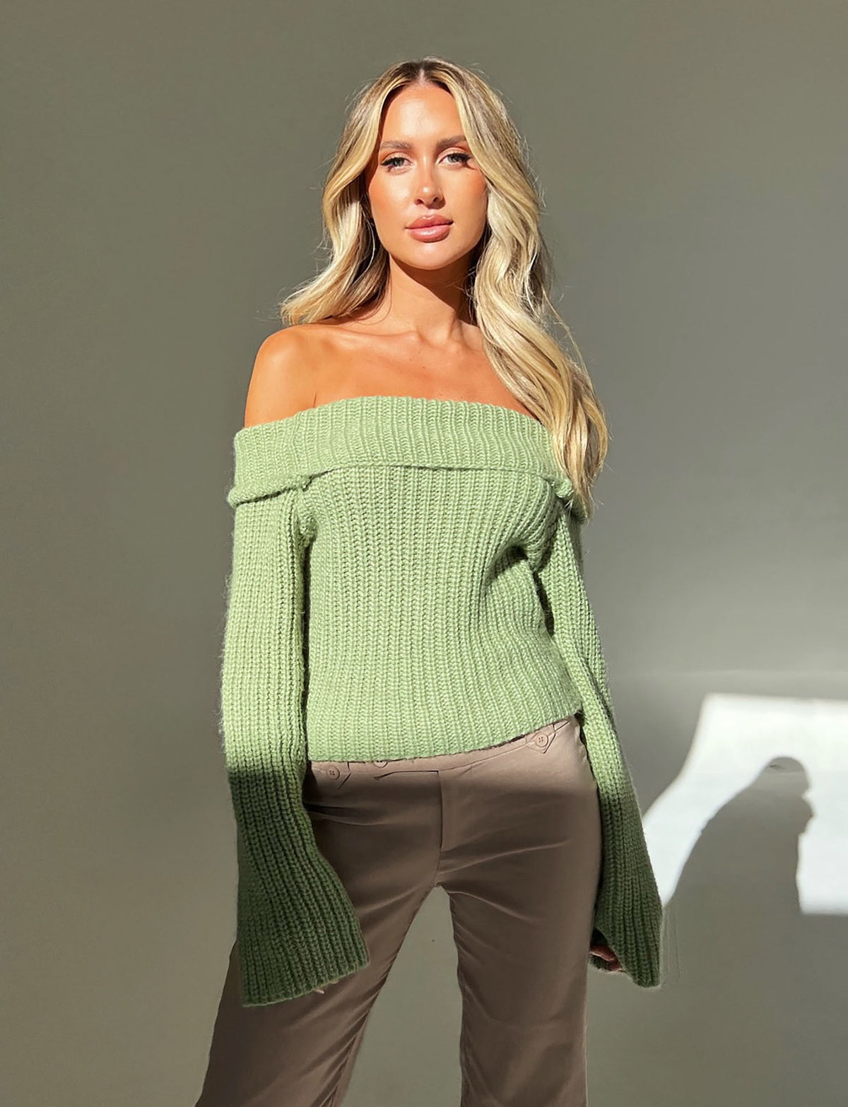 A person with long blonde hair is wearing a chic, off-the-shoulder ASHLYN JUMPER - GREEN from TIGERMIST and beige pants. They are standing against a gray background with sunlight casting a shadow.