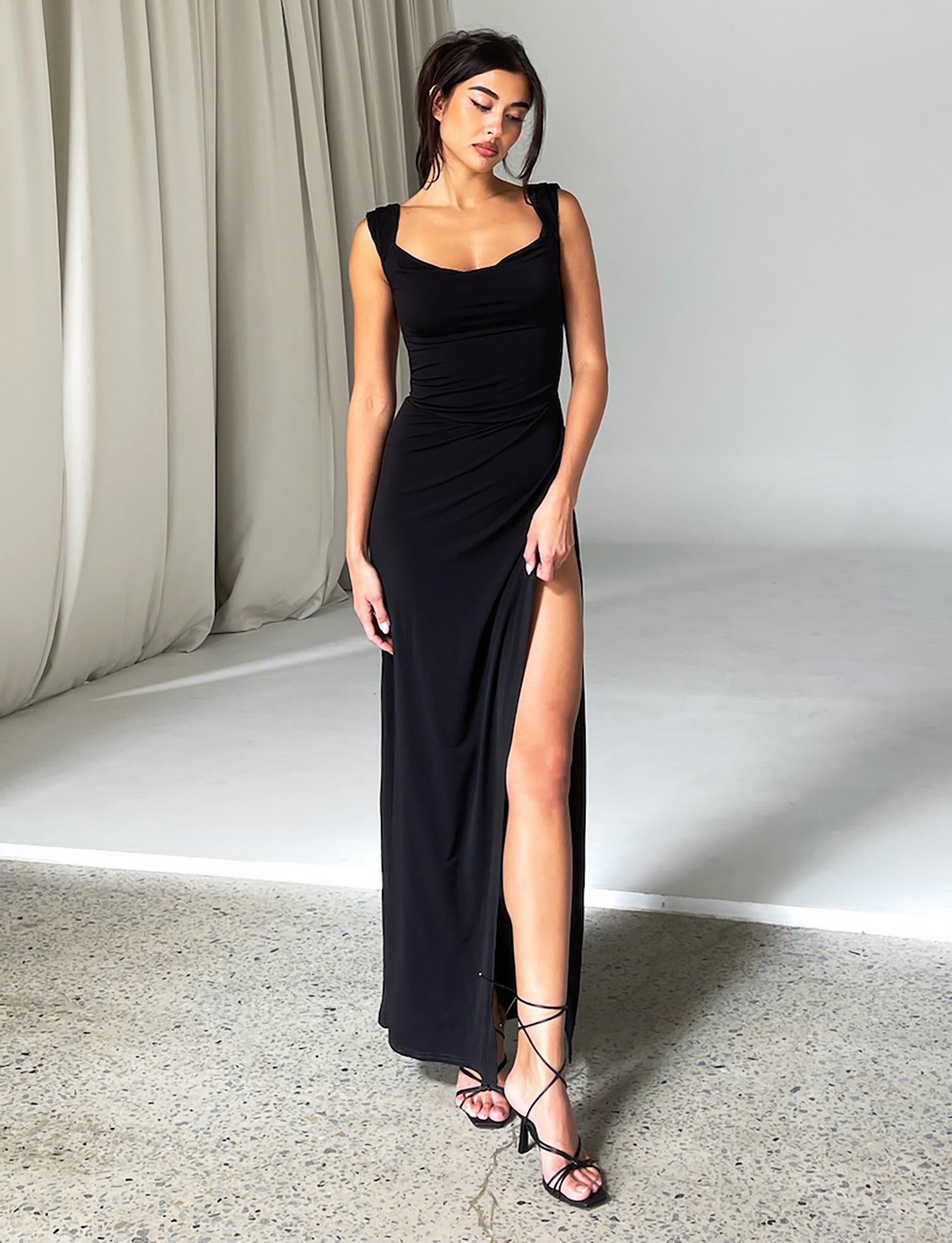 Against a plain backdrop indoors, a woman stands wearing the JOSLIN MAXI DRESS - BLACK from TIGERMIST. The form-fitting, floor-length black dress features a high slit on one side. She completes her look with strappy black high heels. Her dark hair is styled up, and she gazes slightly downward with one hand resting on her hip.