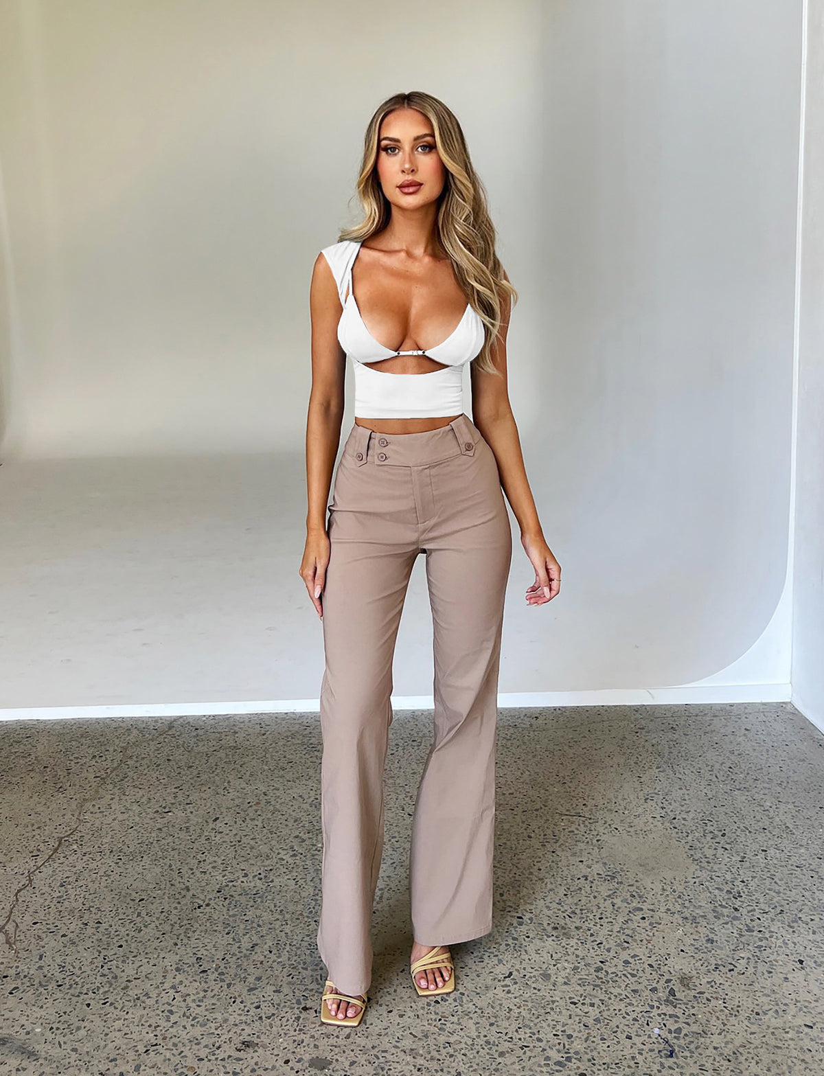 A woman with long hair stands posing against a plain backdrop. She is wearing a white, fitted crop top with cutouts and the KITTIE HIGH RISE PANT - BEIGE : TAN from TIGERMIST. She is also wearing beige open-toe sandals. Her expression is neutral, and her hands are relaxed by her sides.