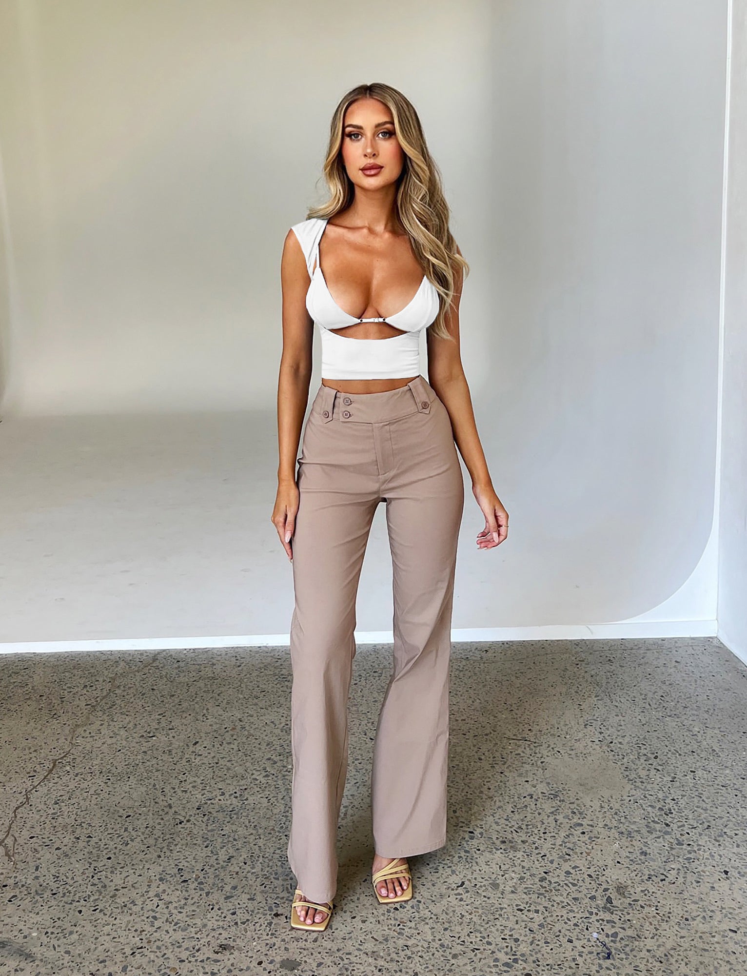 A woman with long hair stands posing against a plain backdrop. She is wearing a white, fitted crop top with cutouts and the KITTIE HIGH RISE PANT - BEIGE : TAN from TIGERMIST. She is also wearing beige open-toe sandals. Her expression is neutral, and her hands are relaxed by her sides.