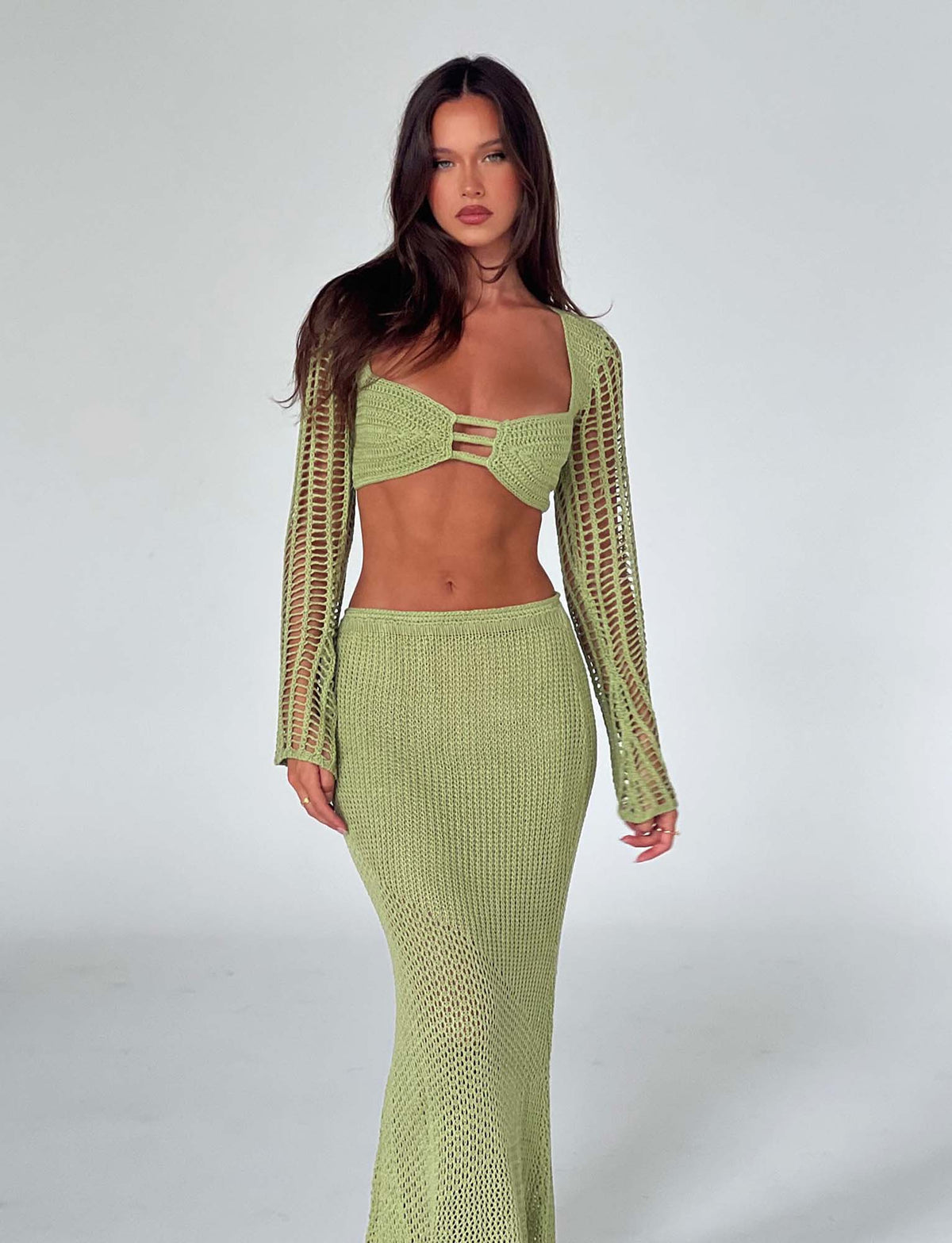 A woman with long dark hair stands confidently against a plain white background. She is wearing the Macauley Top in pastel green from Tiger Mist, a light green crocheted crop top with long sleeves and front cut-out details, paired with a matching floor-length skirt.