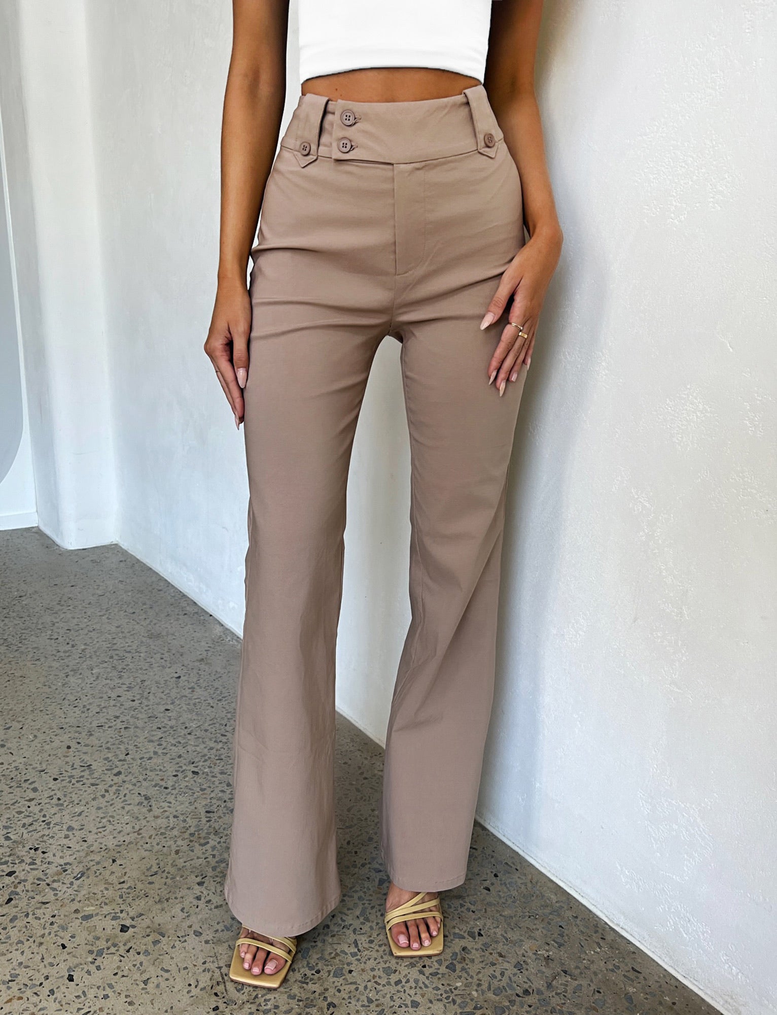 A person is standing indoors against a light-colored wall, exuding TIGERMIST vibes in KITTIE HIGH RISE PANTS - BEIGE : TAN and square-toed mustard-colored heels. They are also wearing a white cropped top. The image is cropped at the shoulders and feet.