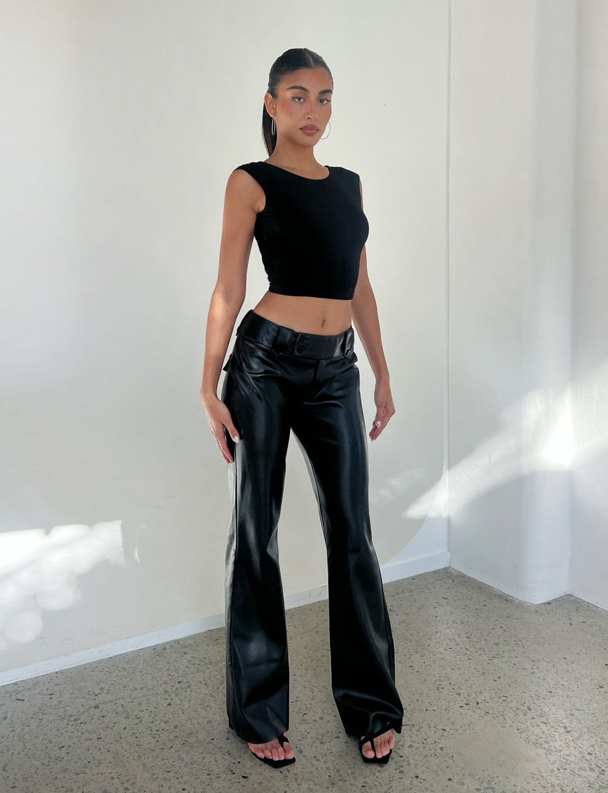 A person with long hair in a ponytail is posing in a brightly lit, minimalistic space. They're wearing the TIGERMIST CHRISTY TOP - BLACK, high-waisted black leather pants, and black open-toe shoes. Their expression is neutral and their hands are by their sides.