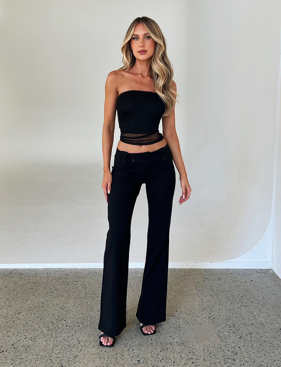 A woman with long, wavy blonde hair stands against a plain white backdrop. She is wearing a TIGERMIST HARRIETT TUBE TOP in black with a sheer panel across the midriff and black high-waisted flared pants. She completes the look with black heeled sandals.