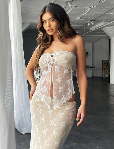 A woman with long, wavy hair stands indoors wearing the sheer, white lace KAROLINA TOP and matching skirt from TIGERMIST. She gazes to the side with a soft expression, as light filters through the sheer curtains behind her into the dimly lit space.