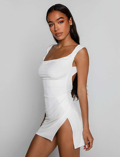 A woman with long, dark hair is wearing the JOSLIN DRESS - WHITE from TIGERMIST, featuring thick shoulder straps, a square neckline, and side slits. She is standing against a plain, light gray background, slightly turned to the side with her head facing the camera.